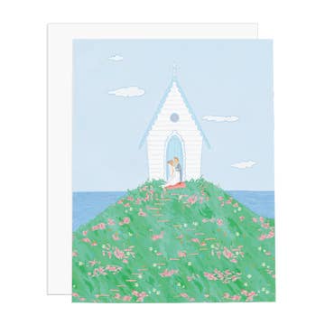 Greeting card with image of a white wedding chapel on a green hill with pink flowers and background of blue sky and water. White envelope included. 