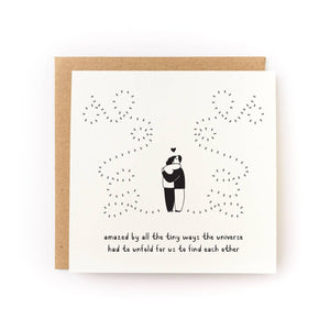 Greeting card with cream background with black drawing of two people hugging with a heart above them and dotted lines to represent their path to each other, Black text says, "Amazed by all the tiny ways the universe had to unfold for us to find each other'. Kraft envelope included. 