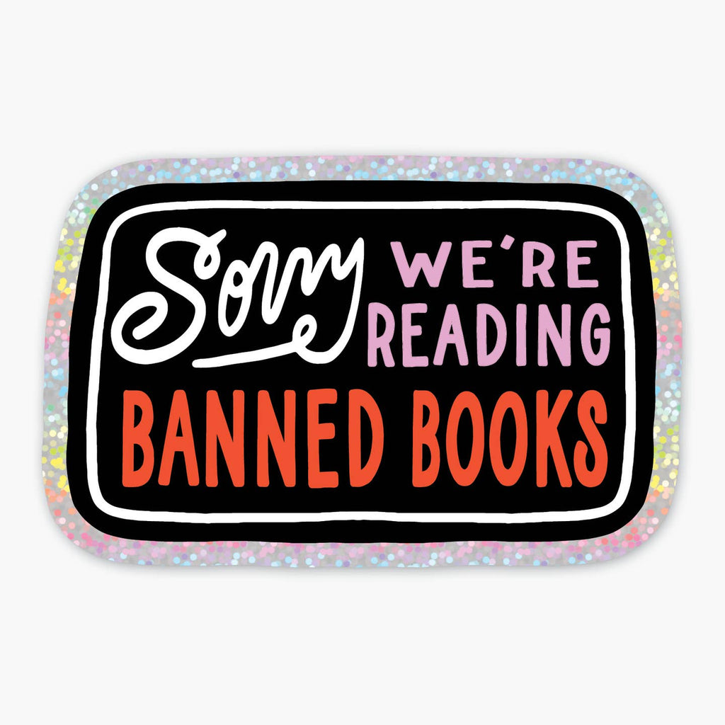 Sticker with black background with iridescent silver border white text says, "Sorry". pink text text says, "We're reading" and red text says, "banned books". 