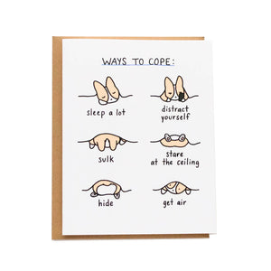 White background with image of tan and white corgi in bed under a blanket in various positions. Black text says, “Ways to cope”, “Sleep a lot”, “distract yourself”, “sulk”. “stare at the ceiling”, “hide” and “get air”.  Kraft envelope included.