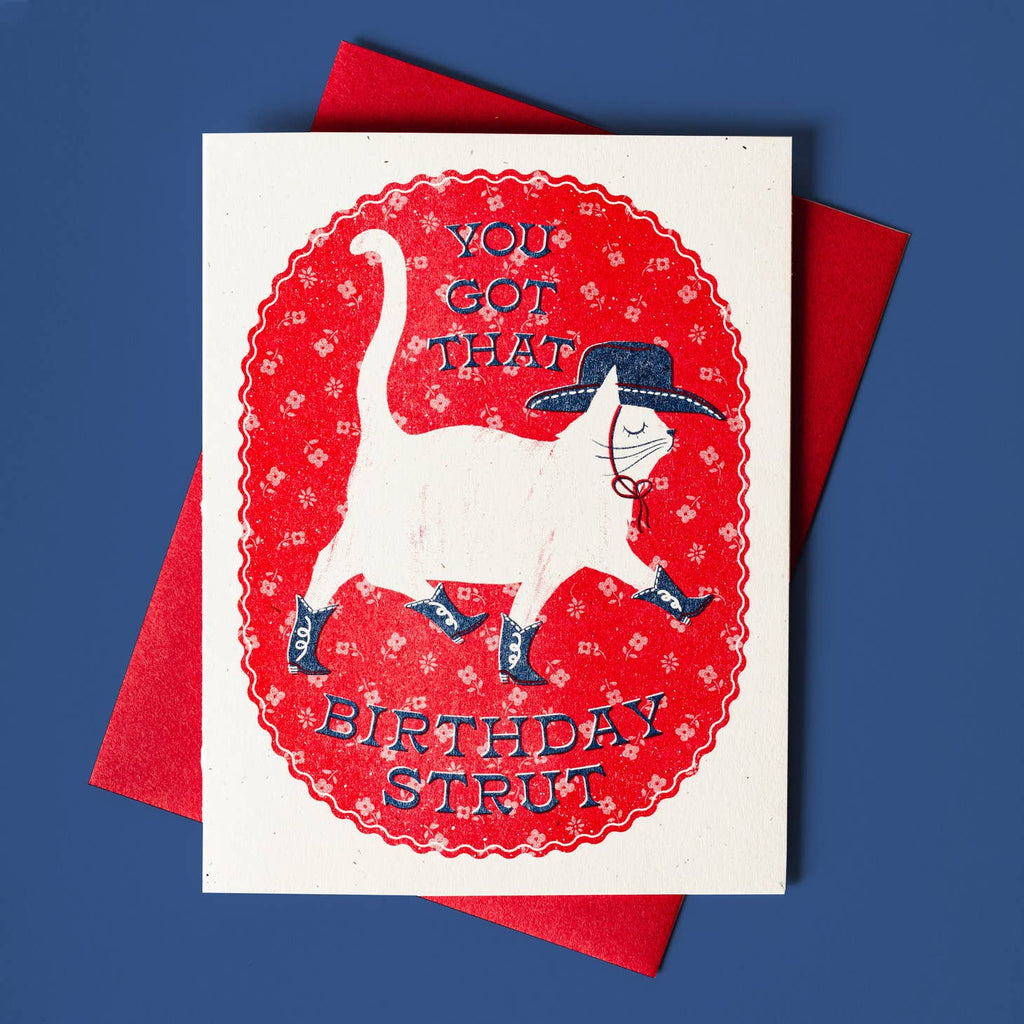 White background with red patterned oval with image of white cat in blue cowboy hat and boots and blue text says, "You got that birthday strut". Red envelope included, 