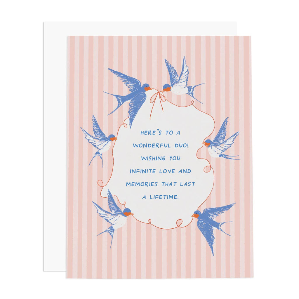 Greeting card with pink and light pink striped background with image of six blue and white swallows holding a red string and tying it in a bow. Blue text says, "Here's to a wonderful duo! Wishing you infinite love and memories that last a  lifetime." White envelope included. 