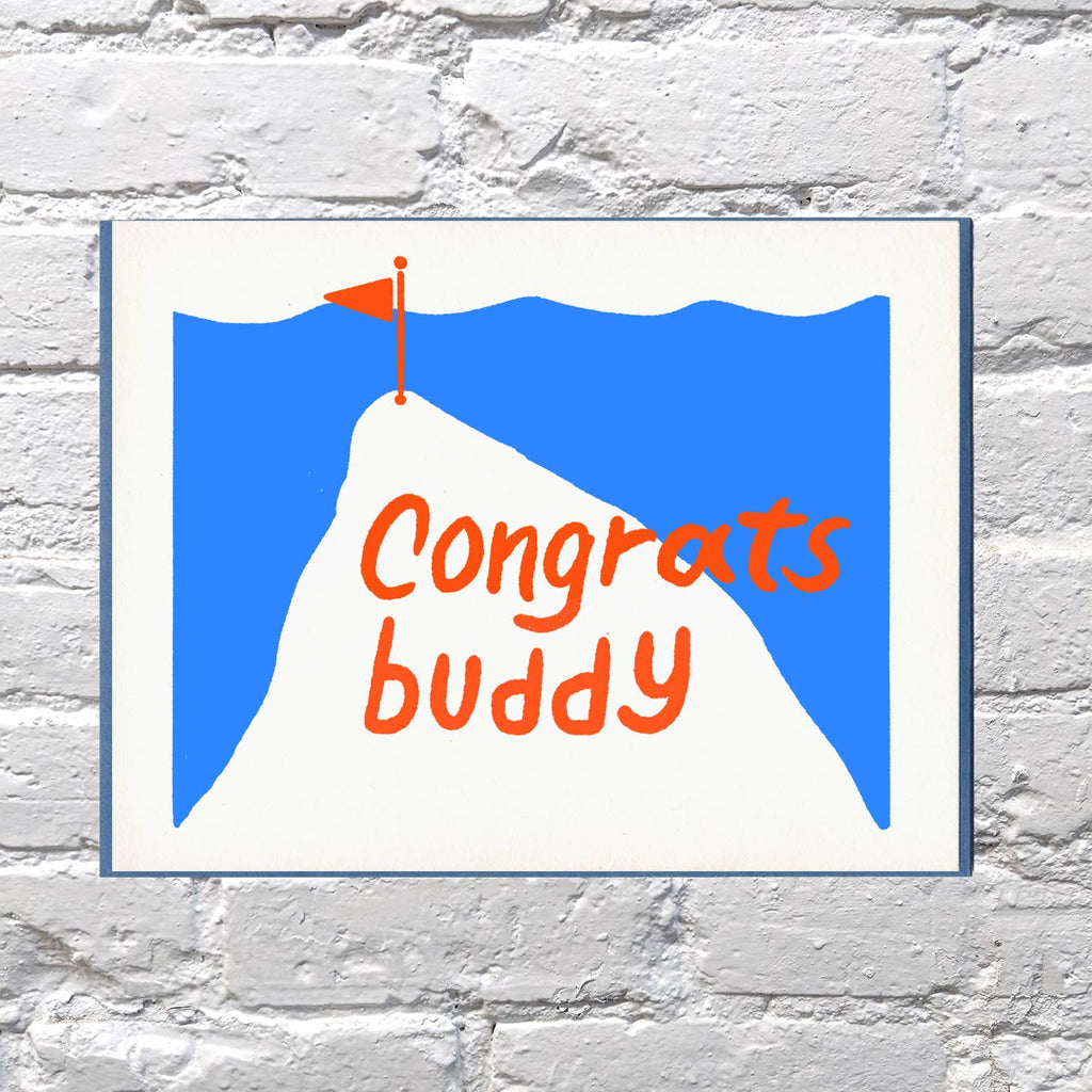 Greeting card with blue and white background with outline of white mountain with a red flag at the top and red text says, "Congrats Buddy". Blue envelope included. 
