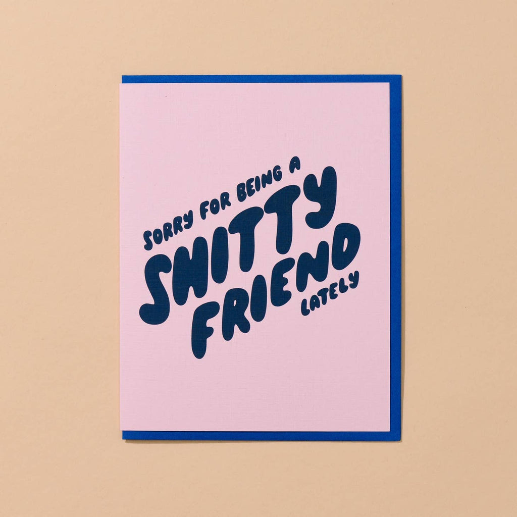 Greeting card with pink background and blue puffy text says, "Sorry for being a shitty friend lately". Blue envelope included. 