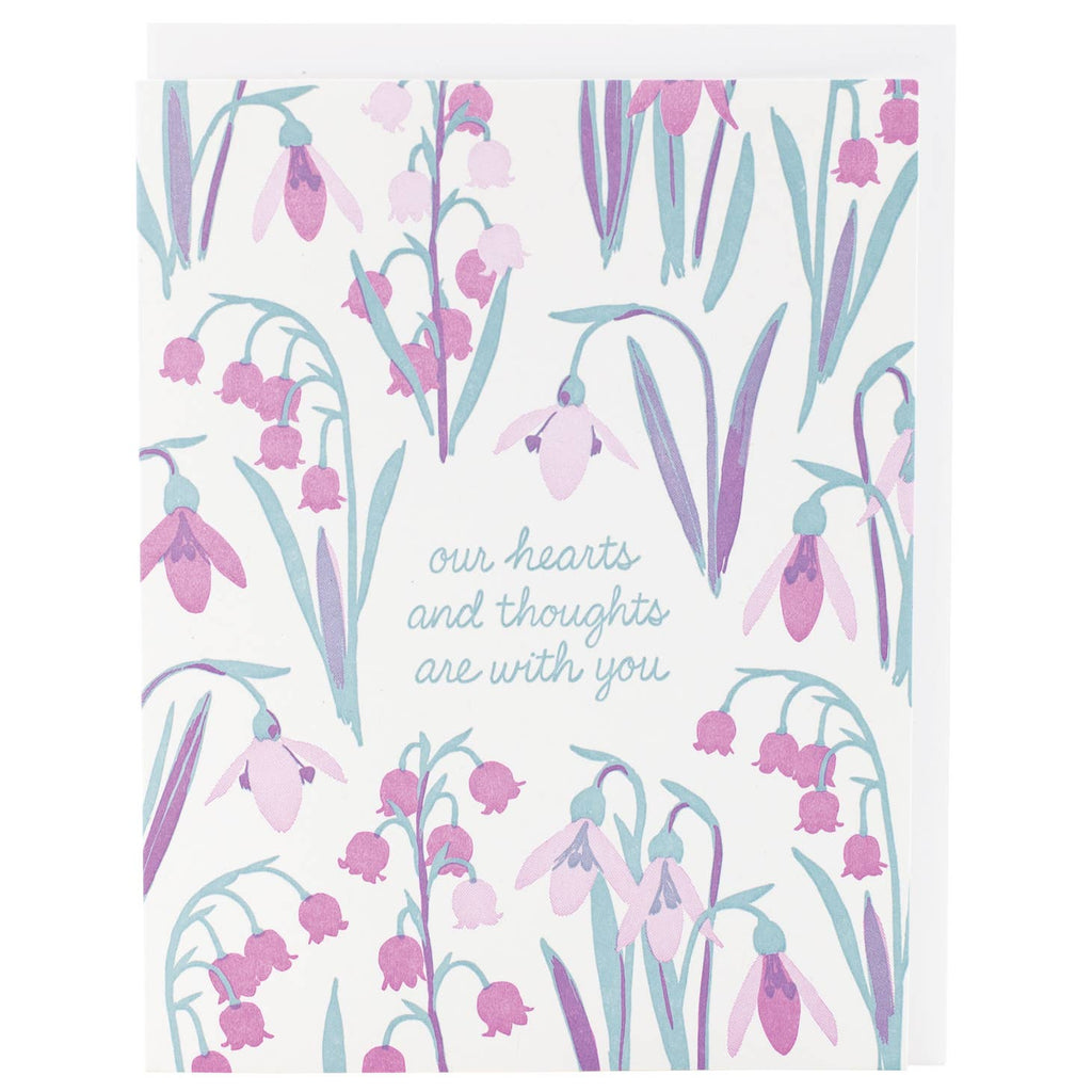 Image of card with white background with images of pink and lavender lily of the valley flowers with green leaves and green text says, “Our hearts and thoughts are with you”. White envelope is included. 