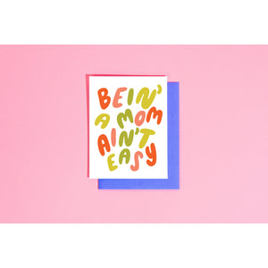 White card with red, pink and green text saying, “Bein’ A Mom Ain’t Easy”. A blue envelope is included.
