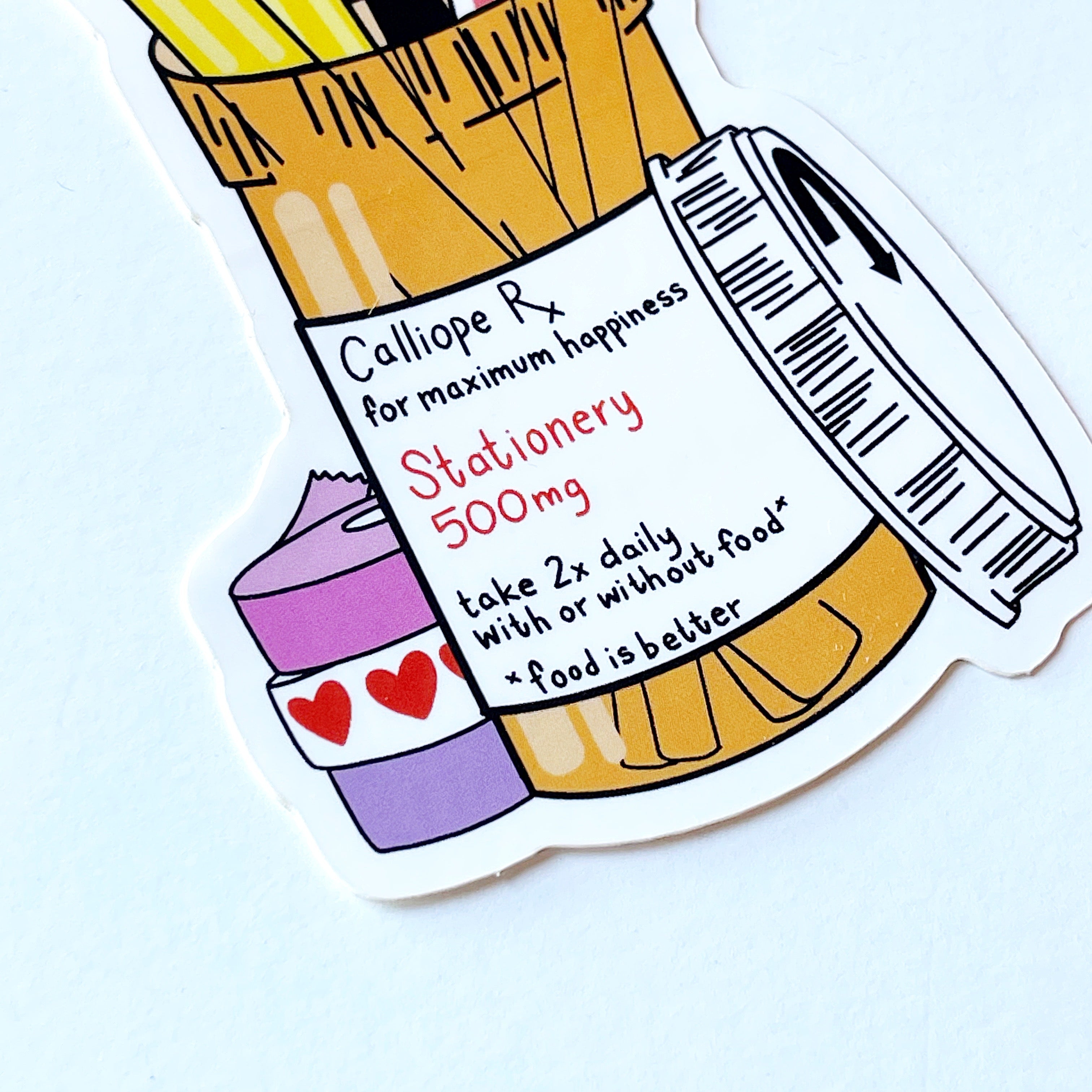 Close up of label: Classic orange prescription bottle with a label that says "Calliope Rx for maximum happiness" in black. Under that "Stationery 500 mg" in red. Under that "take 2x daily with or without food. Food is better."  A stack of 3 rolled tapes are at the bottom left; 1 pink, 1 with red hearts, 1 purple. The cap is leaning on the bottle to the right.