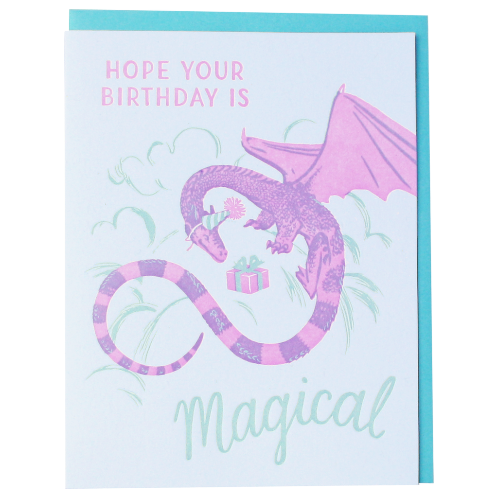 Light blue card with pink and green text saying, “Hope Your Birthday Is Magical”. Image of a purple and pink winged dragon wearing a birthday party hat holding a present. A teal envelope is included.