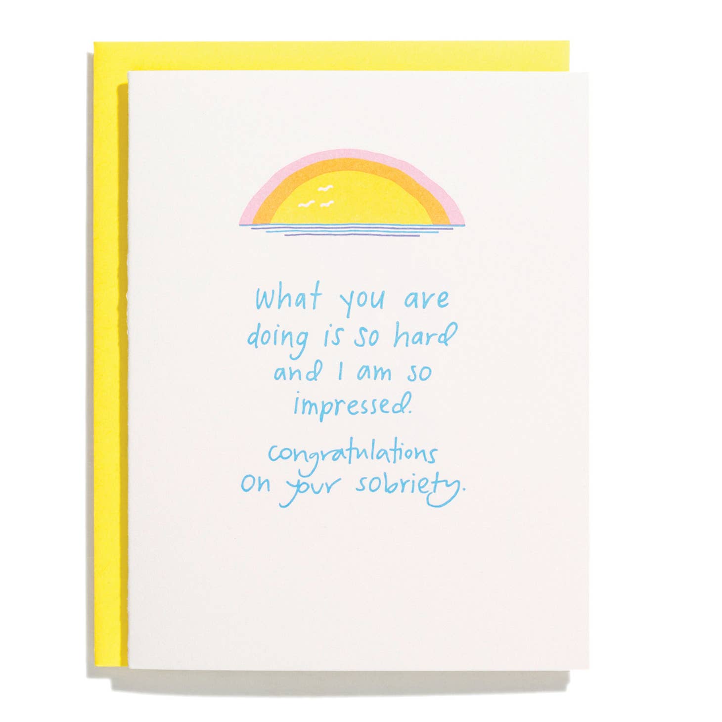 Light pink card with blue text saying, “What you are doing is so hard and I am so impressed. Congratulations on your sobriety.” Images of a yellow, orange and pink sunset over blue water at top of card. A yellow envelope is included.