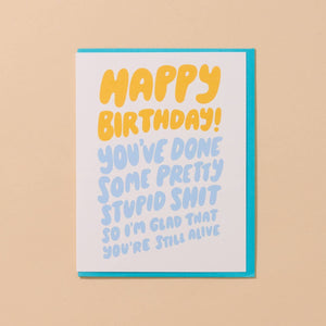 Greeting card with white background with yellow puffy text says, "Happy Birthday!" and blue puffy text says, "You've done some pretty stupid shit so I'm glad that you're still alive". Turquoise envelope included. 