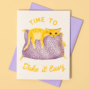 Greeting card with ecru background and image of yellow cat on a purple pillow with yellow text says, "Time to take it easy". Lilac envelope included. 