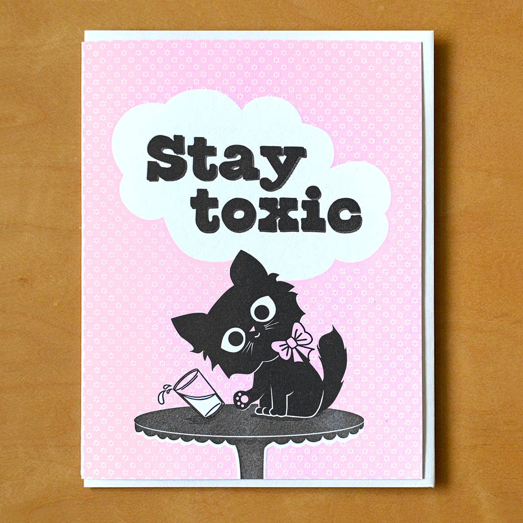 Greeting card with pink background with  image of a black cat with a pink bow knocking over a glass of milk with black text says, "Stay toxic". White envelope included. 