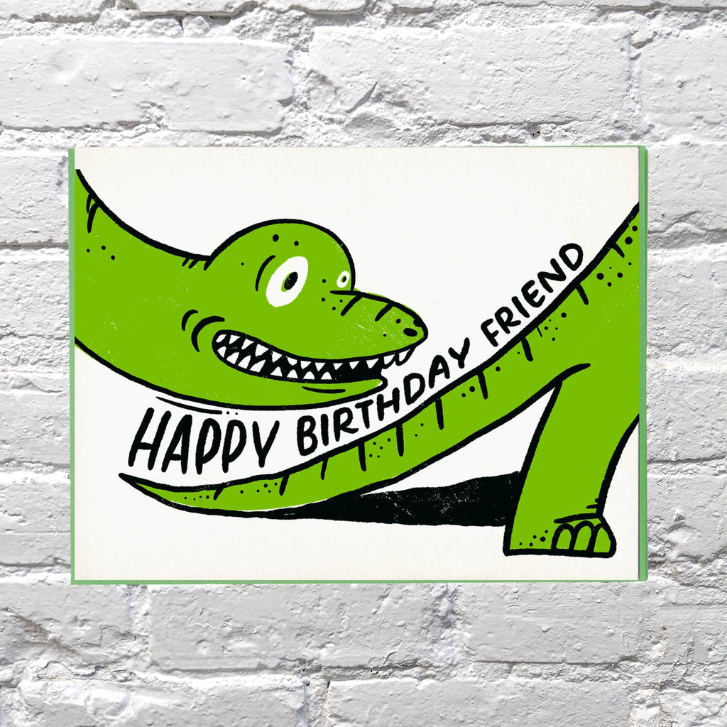Greeting card with white background with image of head and tail of green dinosaur with black text says, "Happy Birthday friend". Green envelope included. 