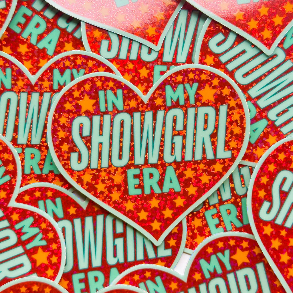 Decorative sticker in heart shape with red background with orange start and aqua text says, "In my showgirl era". 