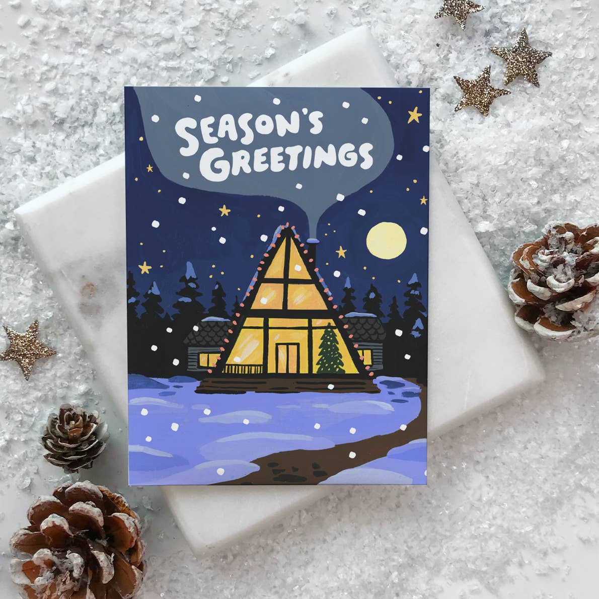 Greeting card with blue background and image of an A-frame house with a tree and holiday lights under a night sky and yellow moon, White text says, "Season's Greetings". Envelope included.