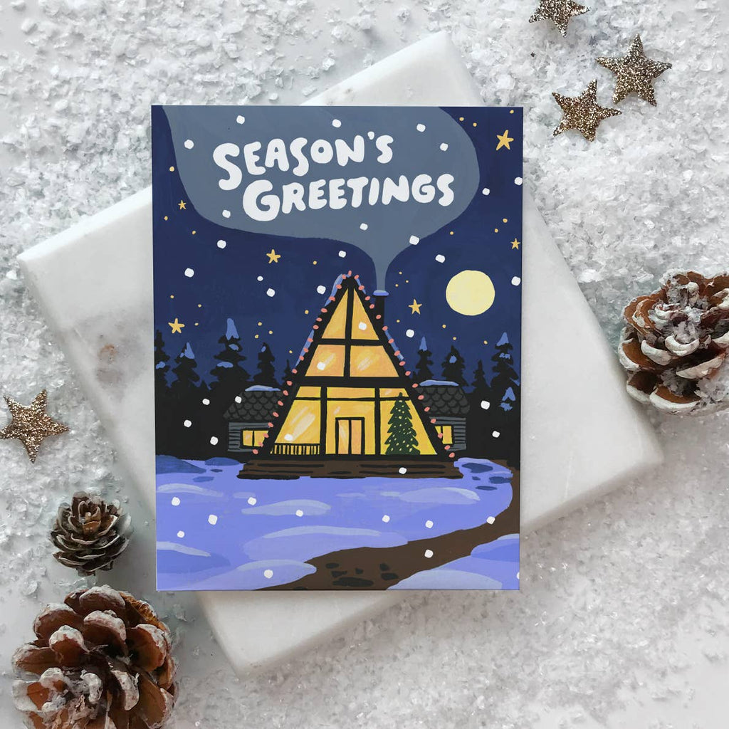 Greeting card with blue background and image of an A-frame house with a tree and holiday lights under a night sky and yellow moon, White text says, "Season's Greetings". Envelope included.