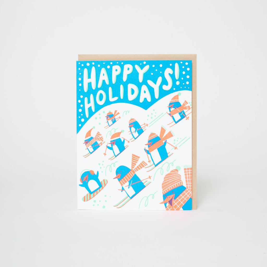 Greeting card with blue and white backgrounds with images of blue and white penguins with peach hats and scarves skiing and white text says, "Happy Holidays!". Kraft envelope included. 