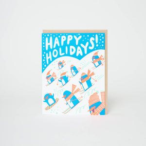 Greeting card with blue and white backgrounds with images of blue and white penguins with peach hats and scarves skiing and white text says, "Happy Holidays!". Kraft envelope included. 