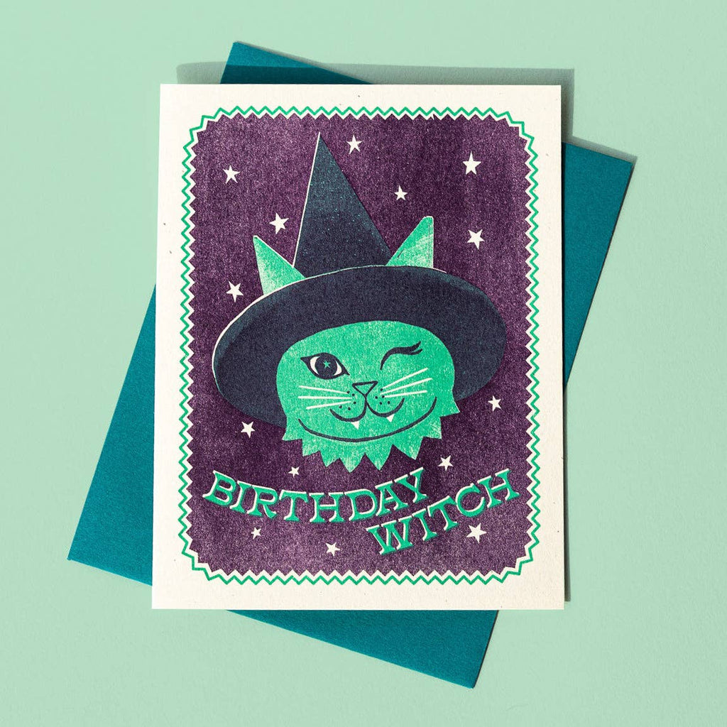 Greeting ard with purple background and white border with neon green zigzag border. Image of green cat wearing a black witch hat winking. Green text says, "Birthday witch". Green envelope included.
