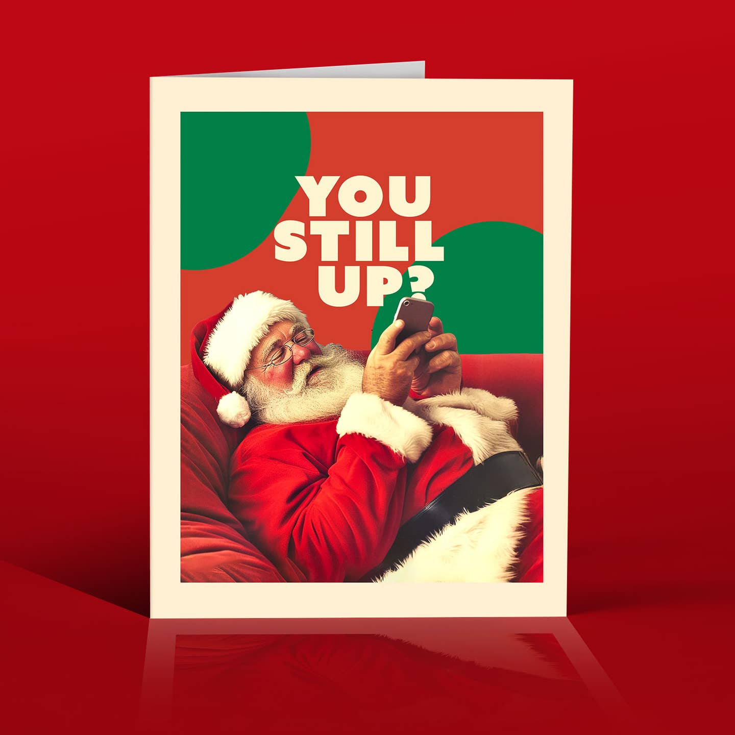 Greeting card with red and green backgrouind with image of Santa reclining on a chair holding a cell phone with white text says, "You still up?". Envelope incuded.