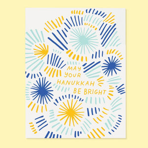 Greeting card with white background and images of bursts and lines in yellow, bright blue and light blue with yellow text says, "May you Hanukkeh be bright". Envelope included.