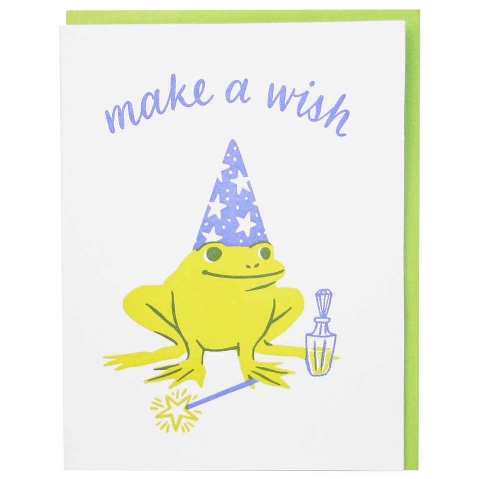 Greeting card with white background and image of a yellow frog wearing a wizard's hat holding a magic wand. Lilac text says, "make a wish". Green envelope included. 