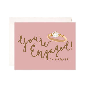 Pink background with image of engagement ring with brown text says, “You’re engaged!”. White envelope included.    