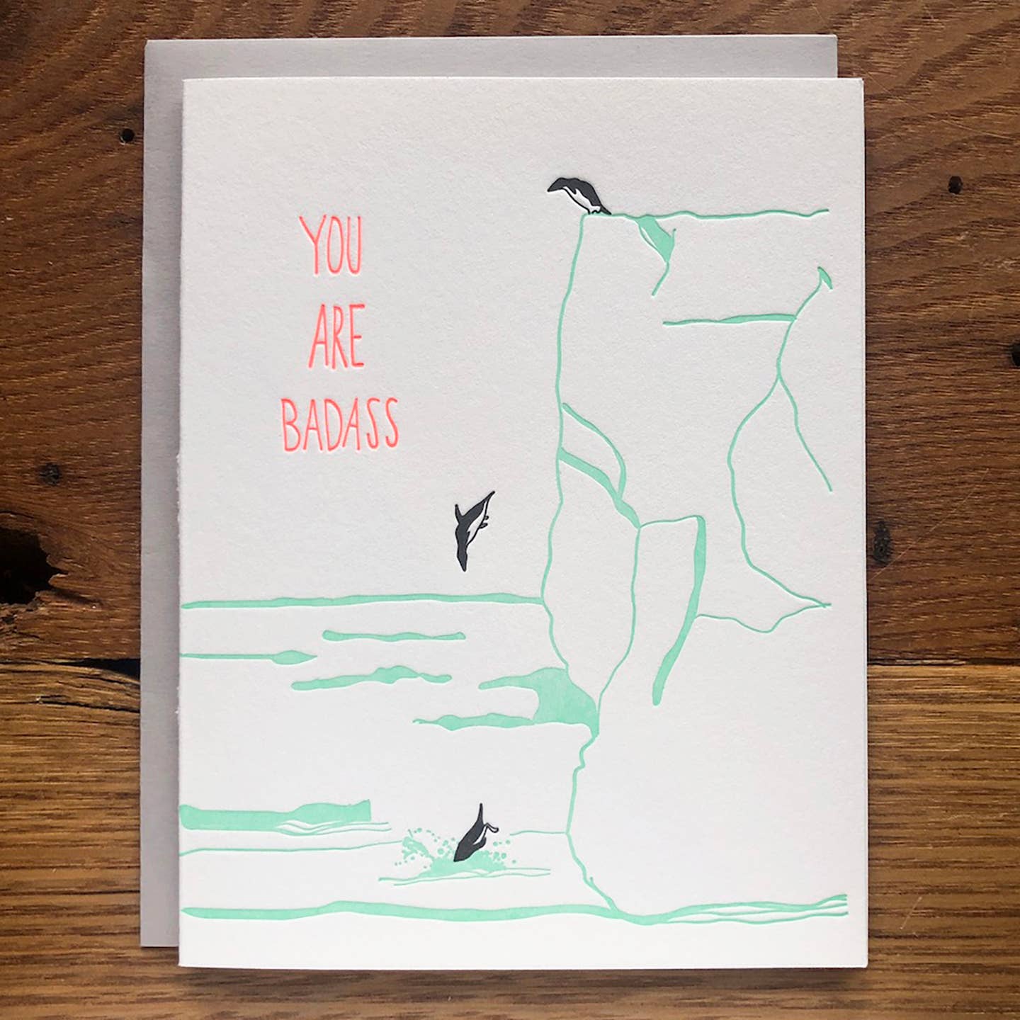 Greeting card with white background and image of black and white penguins jumping off of an aqua cliff into water with pink text says, "You are badass". Envelope included. 