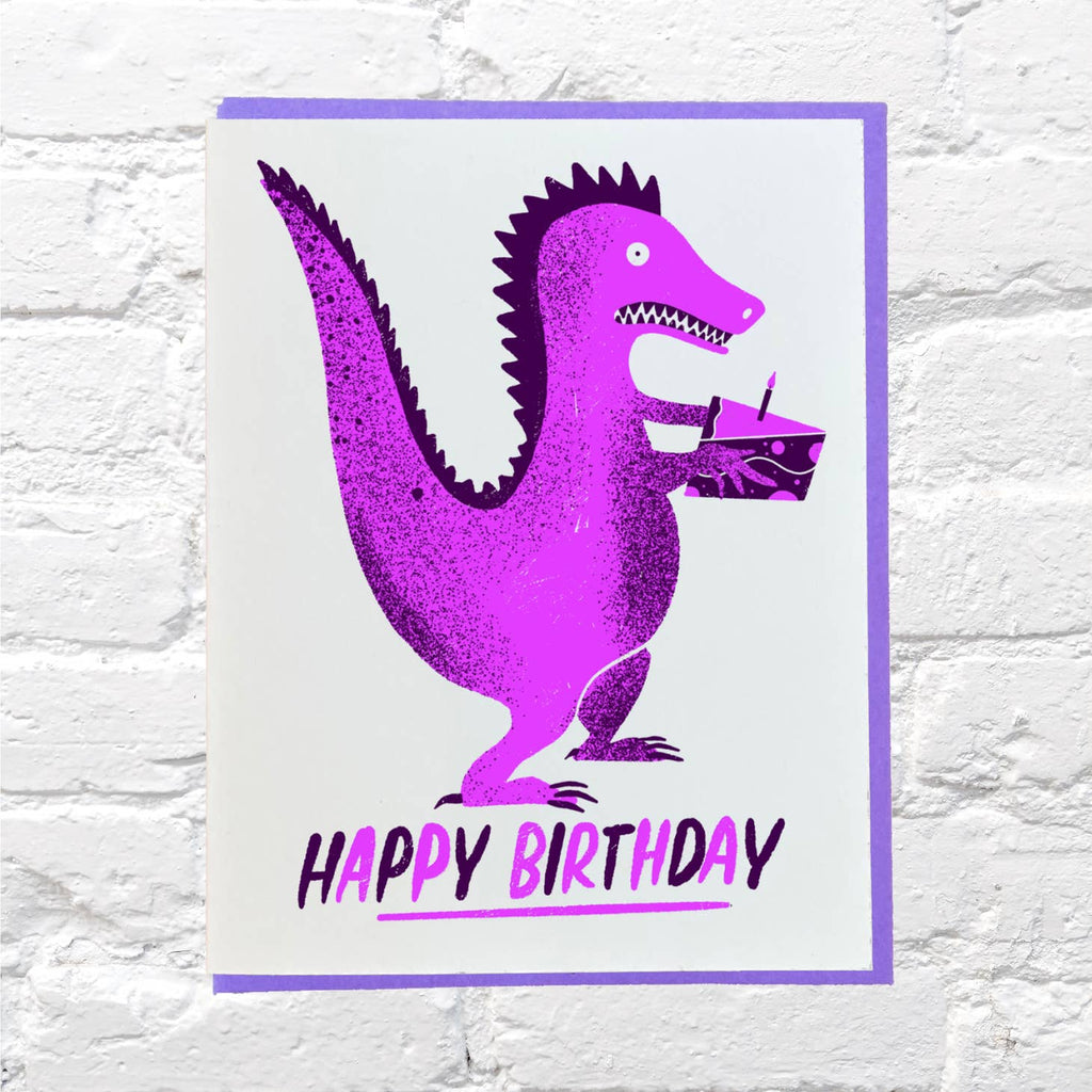 Greeting card with white background and image of purple dinosaur carrying birthday cake. Purple text says, "Happy Birthday". Purple envelope included. 