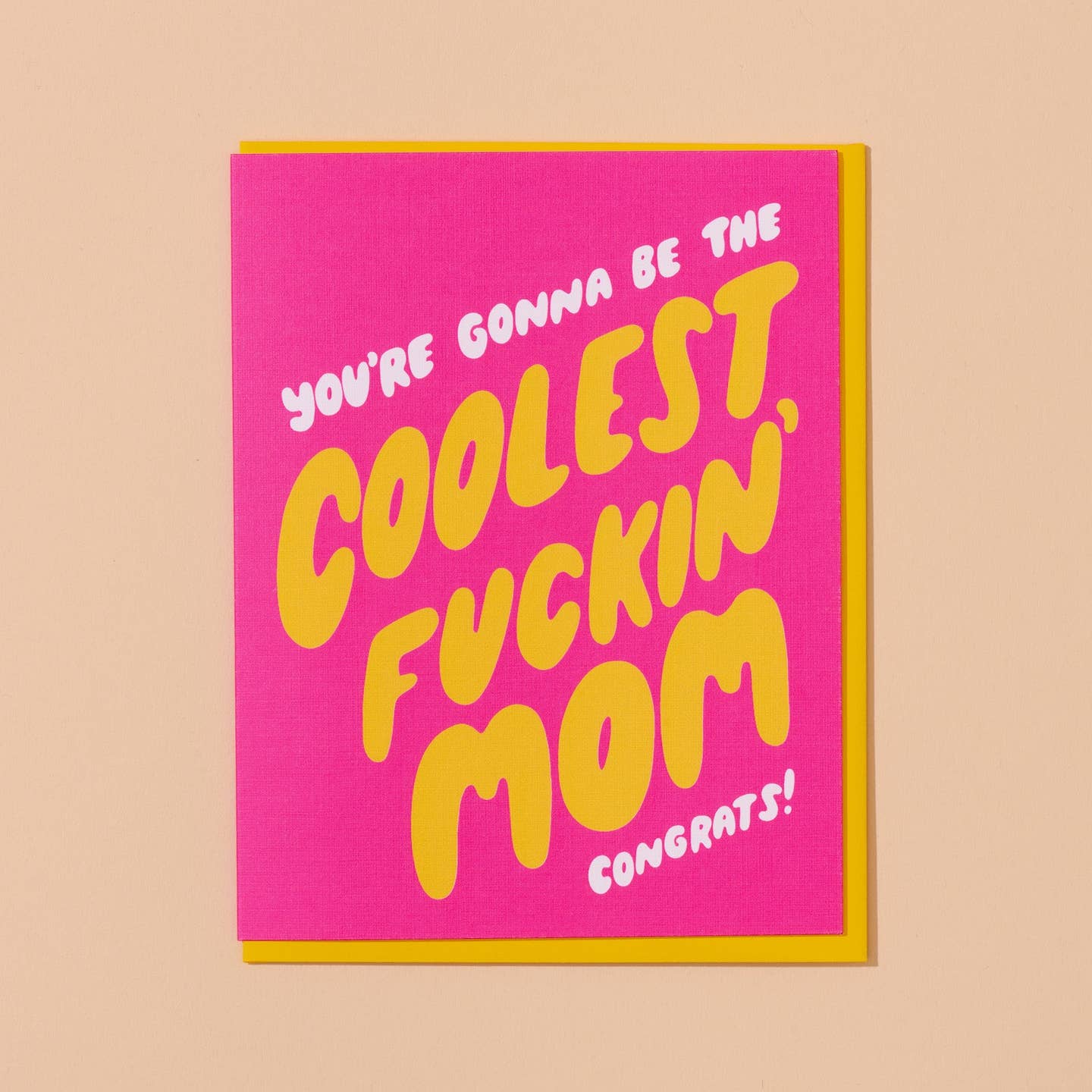 Greeting card with hot pink background with white text says, "You're gonna be the" and yellow text says, "Coolest Fuckin' mom" and white text says, "Congrats!".  Yellow envelope included. 