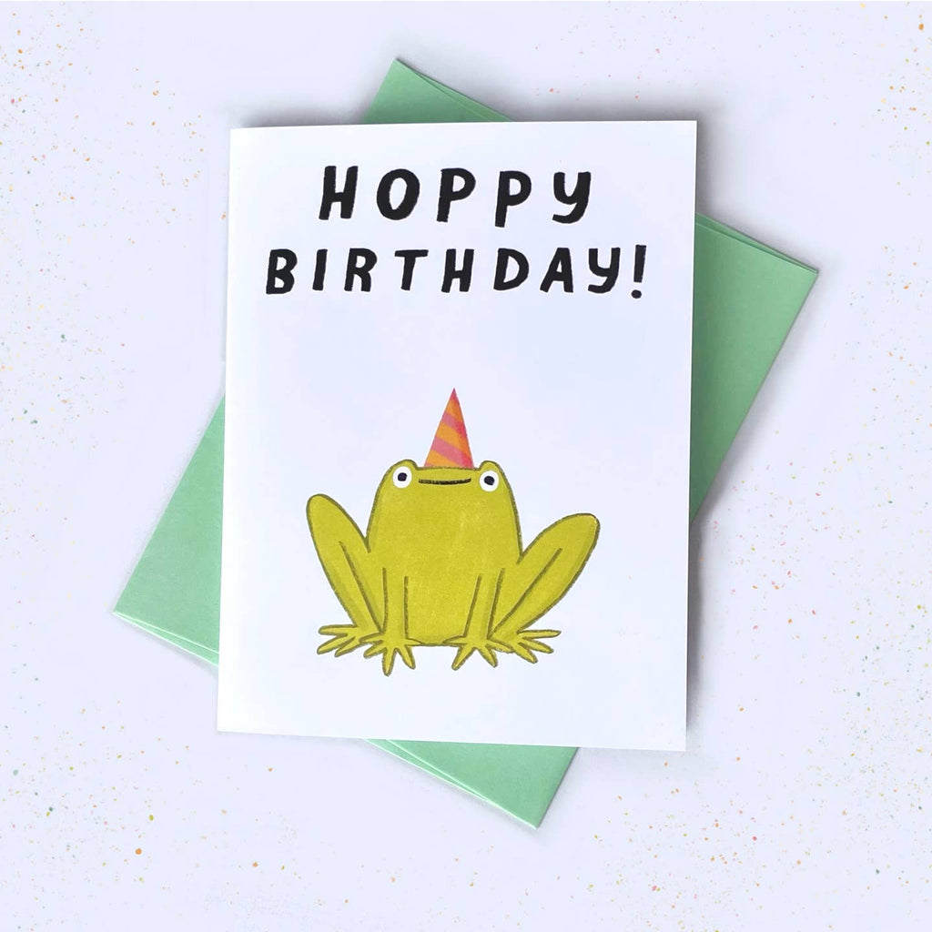 Greeting card with white background and image of a green frog wearing an orange and pink party hat. Black text says Hoppy Brthday!". Green envelope included. 