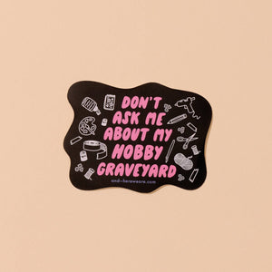 Decorative sticker with black background and white outlined images of hobbies. Pink text says, "Don't ask me about my hobby graveyard". 