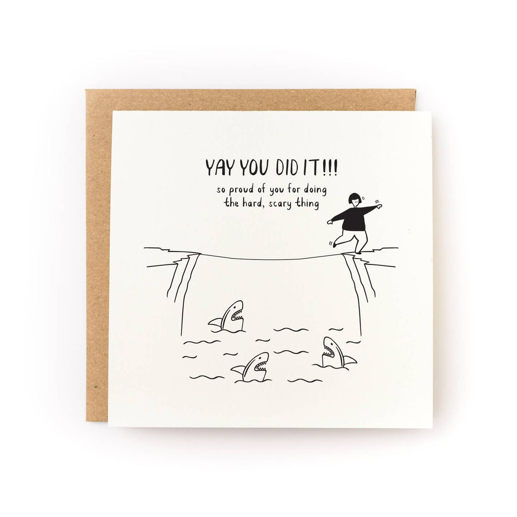 Greeting card with ivory background and black outlined drawing of a person walking on a tight rope over a group of sharks. Black text says, "Yay you did it!!! so proud of you for doing the hard, scary thing". Kraft envelope included. 