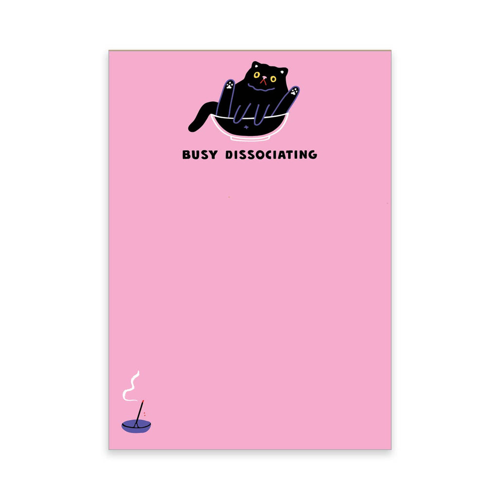 Notepad with pink background and image of a black cat sitting in a bowl with black text says, "Busy dissociating". 