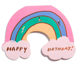 Greeting card in the shape of a rainbow with clouds at each end and gold foil text says, "Happy Birthday". Images of three women sliding down the rainbow. Envelope included. 