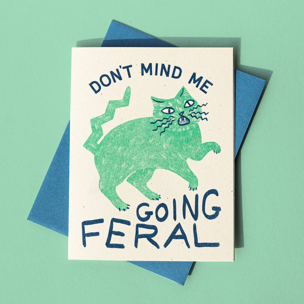 Greeting card with cream background with image of green cat looking wild with blue text says, "Don't mind me going feral". Blue envelope included. 