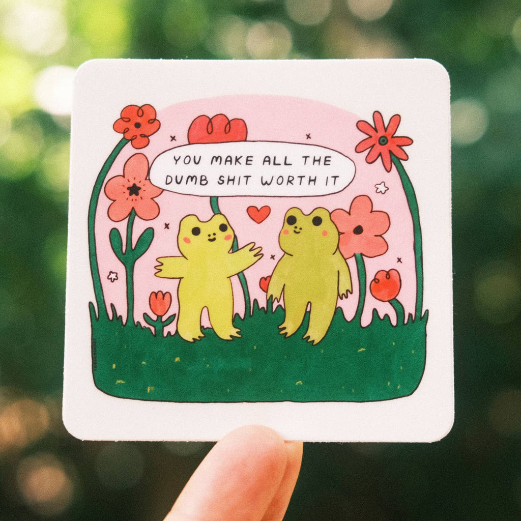 Sticker with pink background and images of two green frogs standing on a field of green grass with red flowers and black text says,"You make all the dumb shit worth it". 
