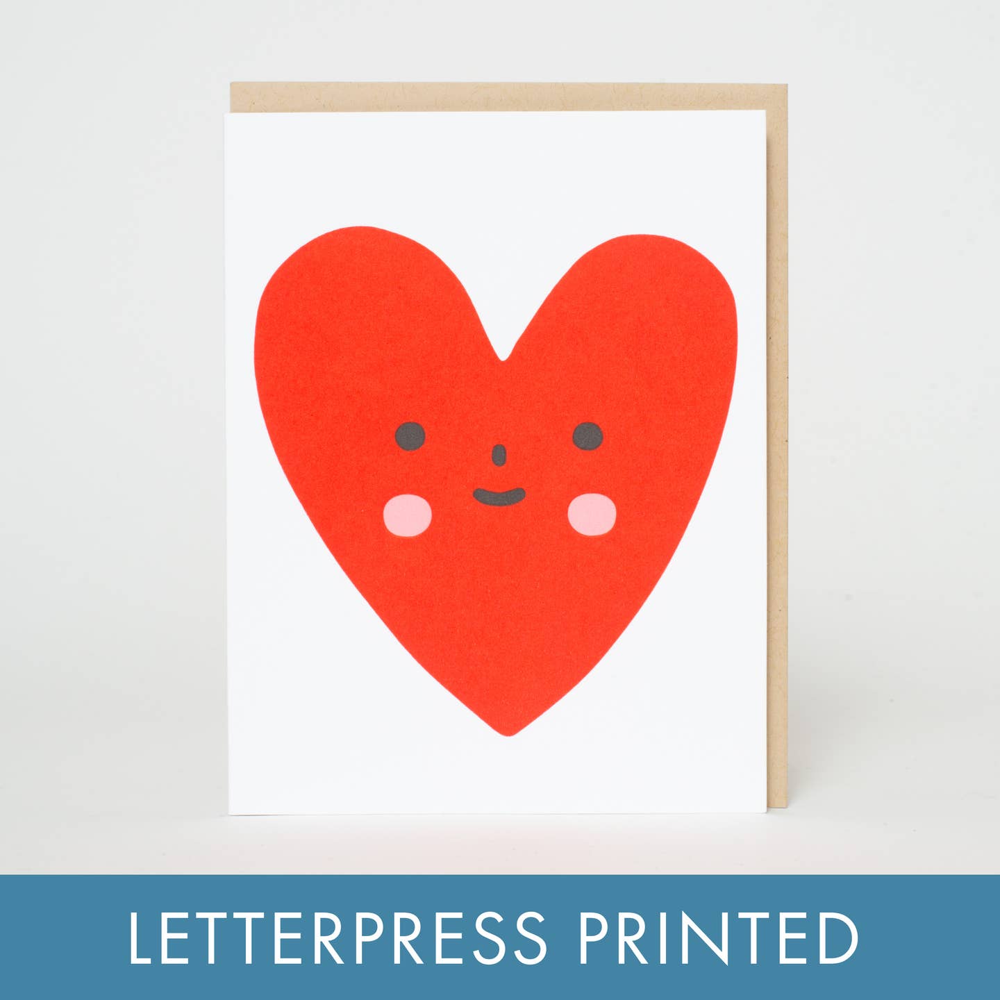 Greeting card with white background and image of red heart with pink cheeks and black eyes, nose and mouth. Kraft envelope included. 