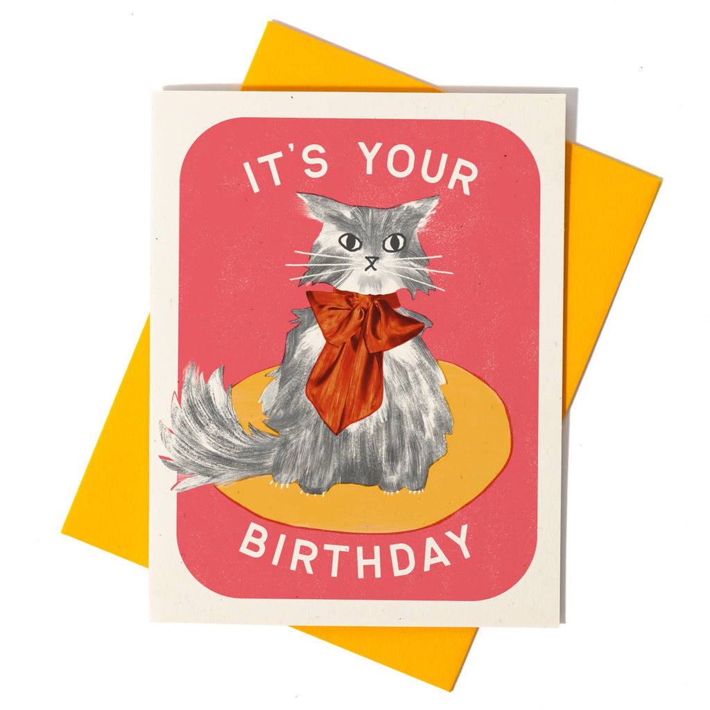Greeting card with light red background with cream border and image of a grey and white cat wearing a red bow. Cream text says, "It's your birthday". Gold envelope included.