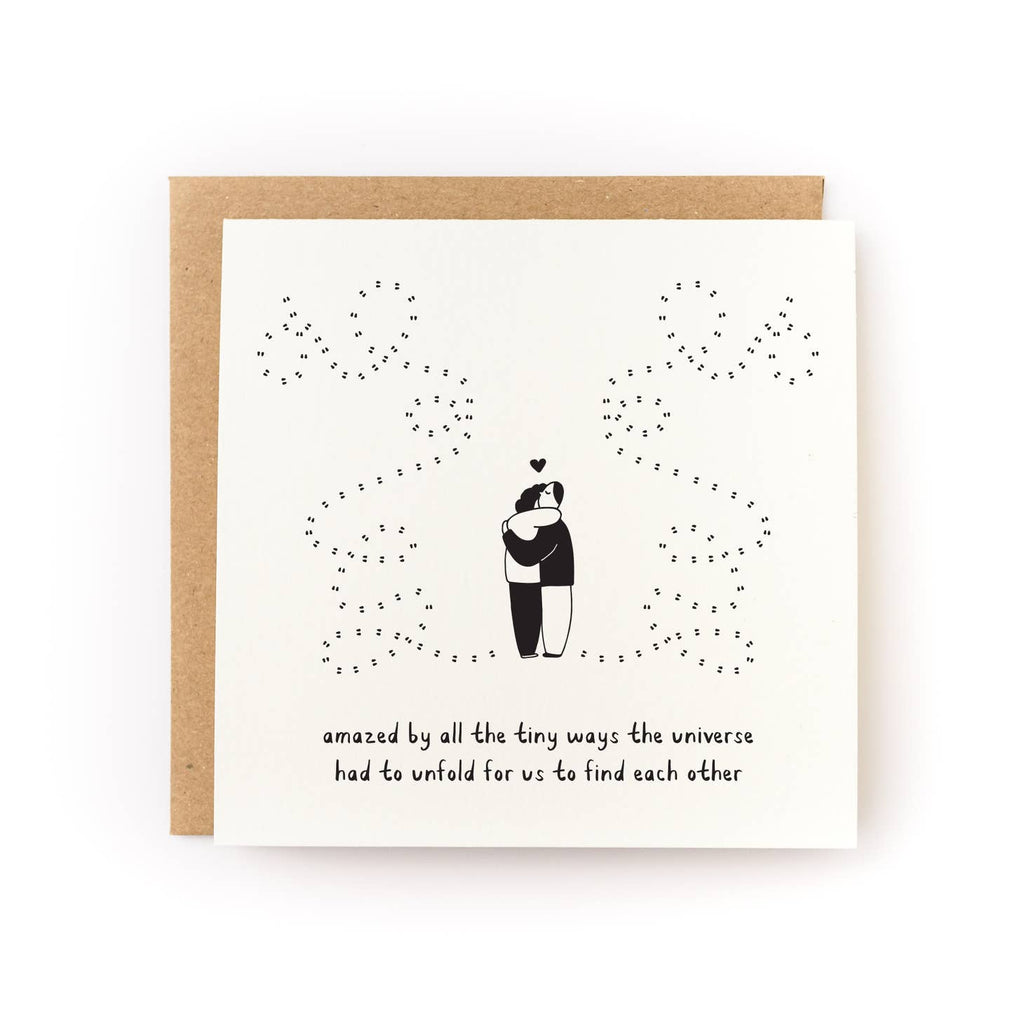 Greeting card with cream background with black drawing of two people hugging with a heart above them and dotted lines to represent their path to each other, Black text says, "Amazed by all the tiny ways the universe had to unfold for us to find each other'. Kraft envelope included. 