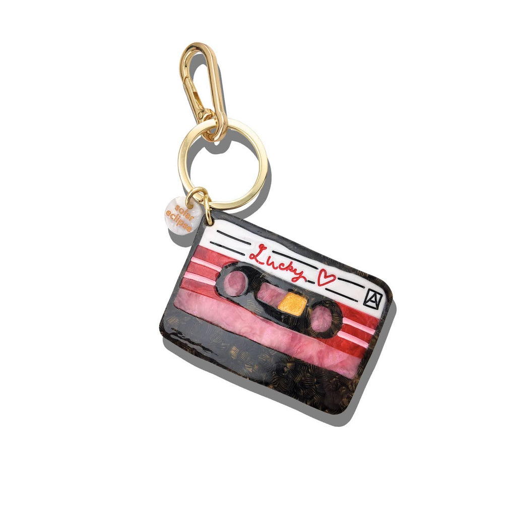 Image of a keychain charm in the shape of a cassette tape with red text says, "Lucky" with a small red heart. Gold colored keyring and clip.