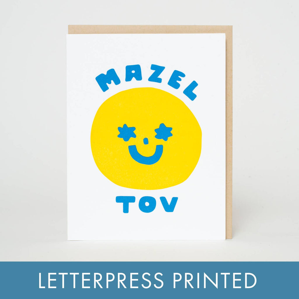 Greeting card with white background with yellow face with blue star eyes, nose and mouth. Blue text says, "Mazel Tov". Kraft envelope included. 