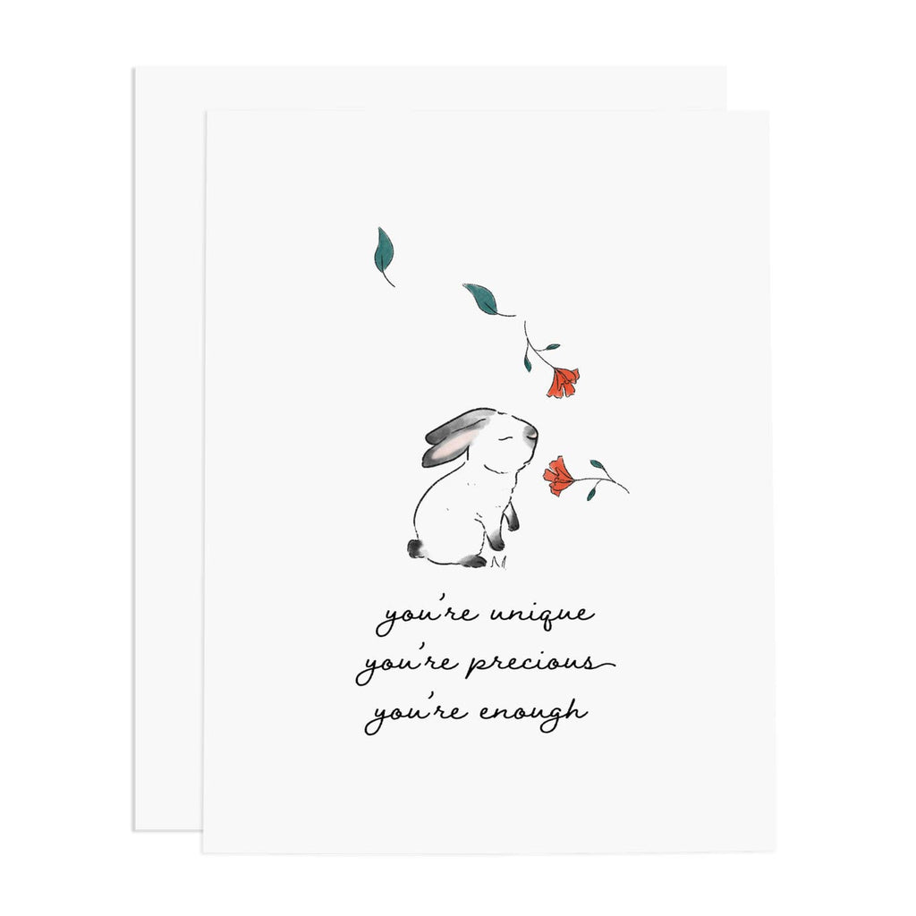 Greeting card with white background and image of a bunny with red flowers falling down in front of it. Black text says "You're unique, you're precious, you're enough". White envelope included.