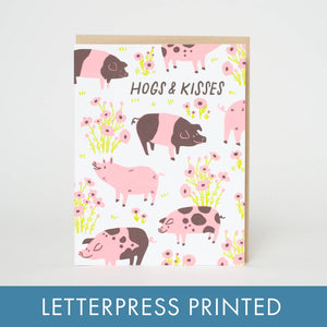 Greeting card with white background and images of brown and pink pigs and pink and brown flowers with brown text says, "Hogs & kisses". Kraft envelope included. 