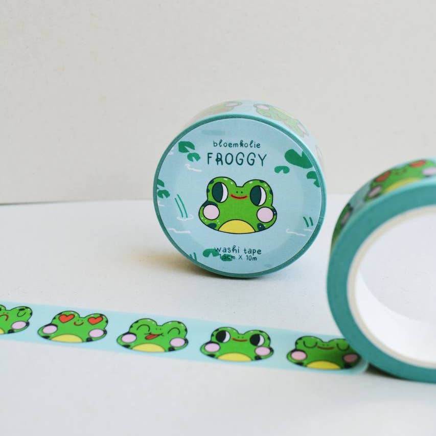 Image of roll of washi tape with aqua background and images of frog faces in green and yellow with various expressions. 