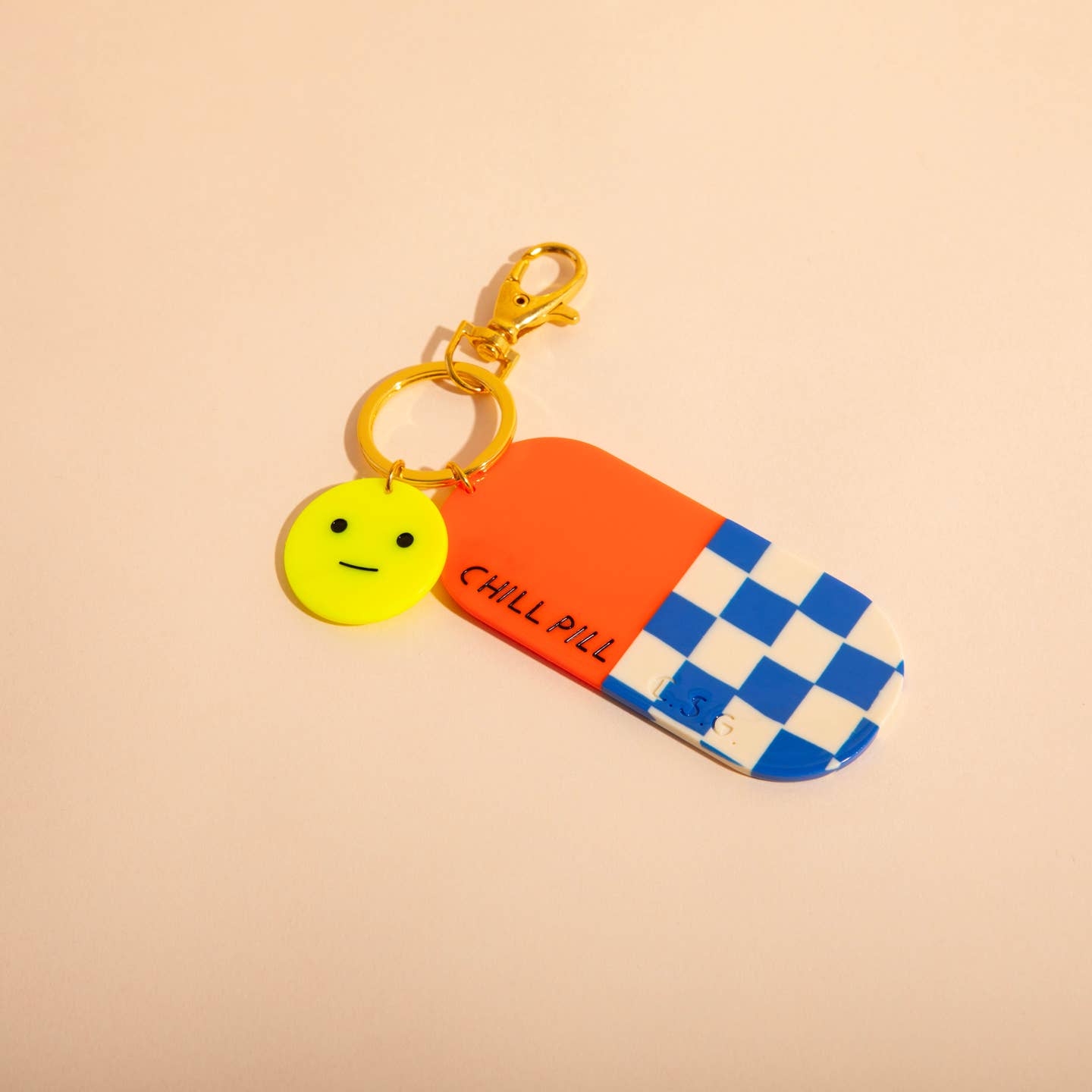 Image of a plastic charm in the shape of a pill capsule in red and blue and white checkerboard and a yellow chill face. 