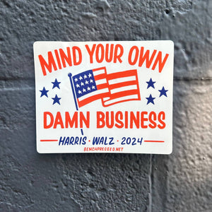 Decorative sticker with white background and image of an American flag and stars in red and blue with red text says, "Mind your own damn business" and blue text says, "Harris Walz 2024". 