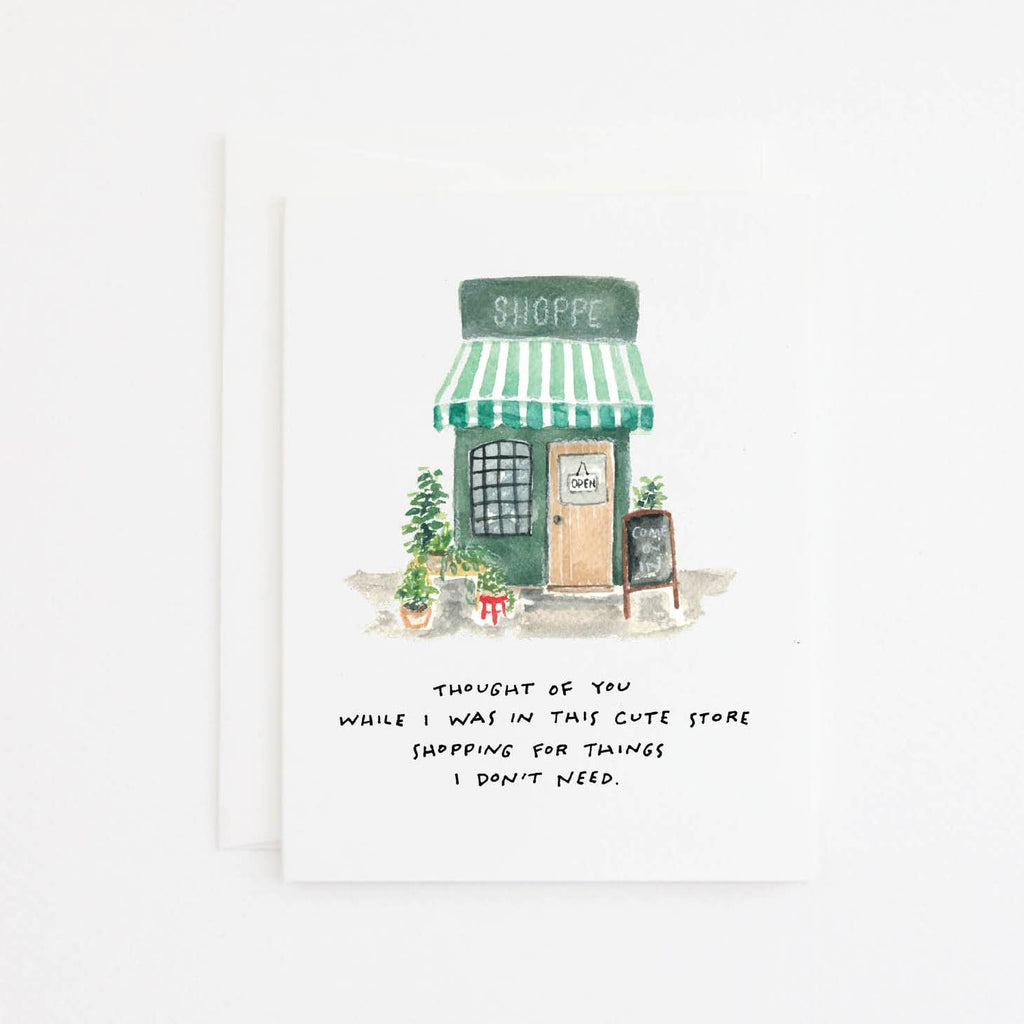 Greeting card with white background and image of small green shoppe with black text says, "Thought of you while I was in this cute store shopping for things I don't need.". White envelope included. 