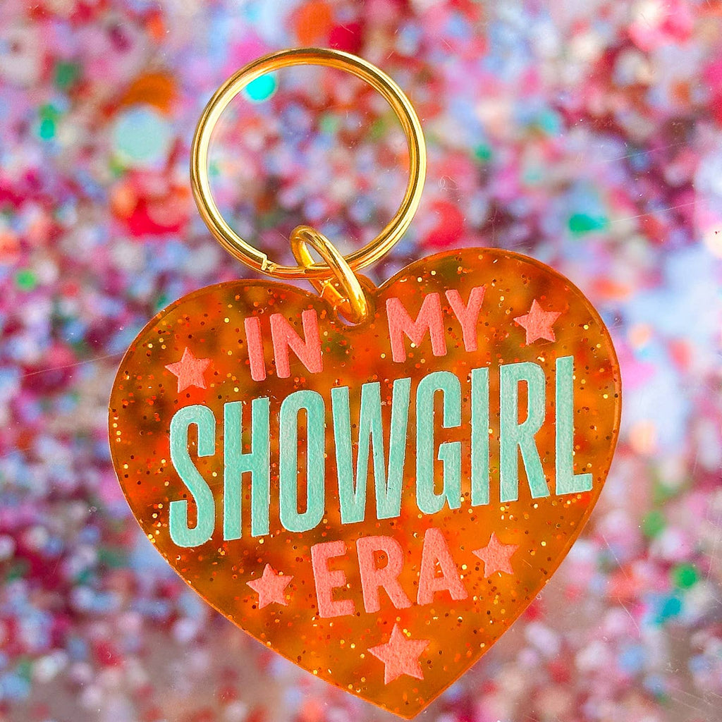 Image of a keychain in gthe shape of a heart with red background and pink stars Pink and aqua text says, "In my showgirl era". 