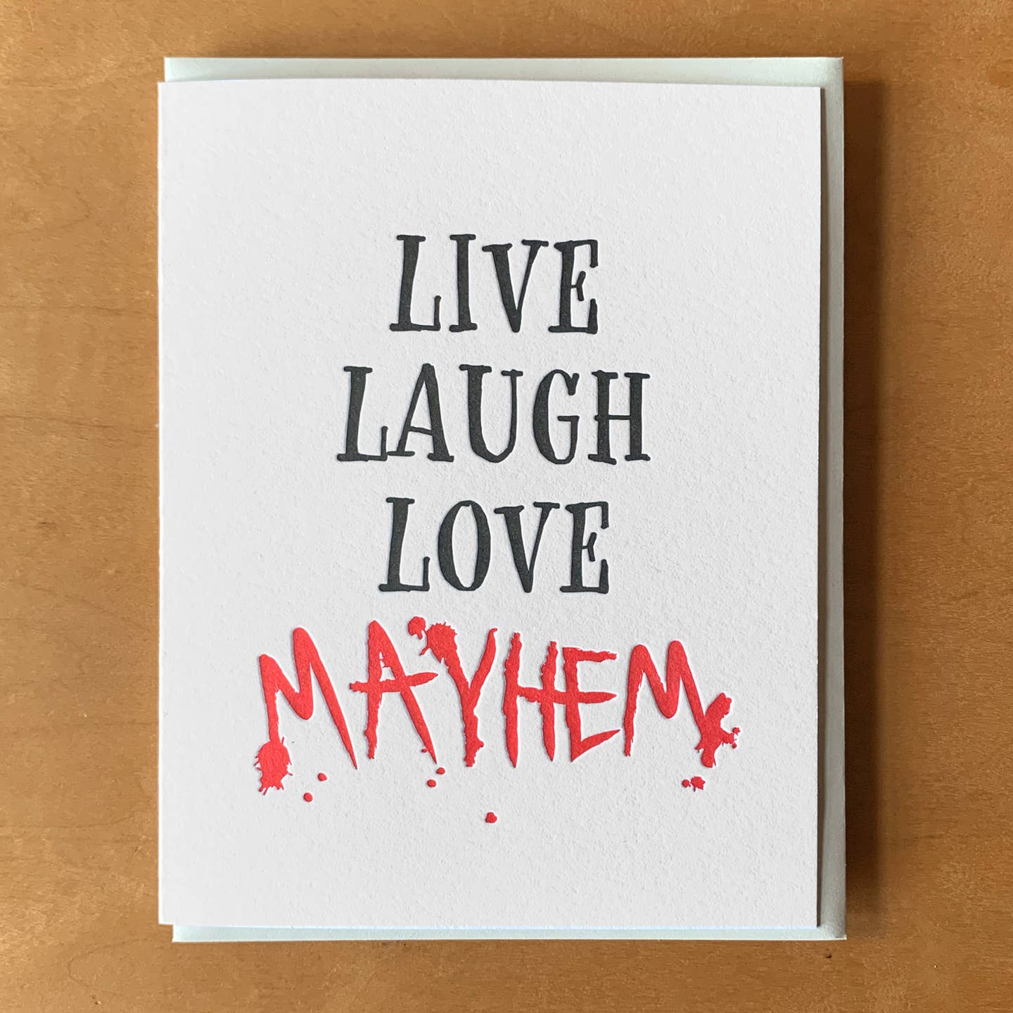 Greeting card with white background with black text says, "Live Laugh Love" and red text says, "Mayhem". Tan envelope included.